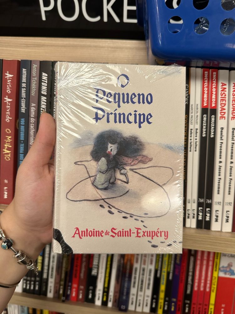 Mão segurando um livro de capa dura com o texto “O Pequeno Príncipe” em azul na parte de cima e “Antoine de Saint Exupéry” em vermelho na parte de baixo e um desenho de uma criança desenhando um avião na areia.