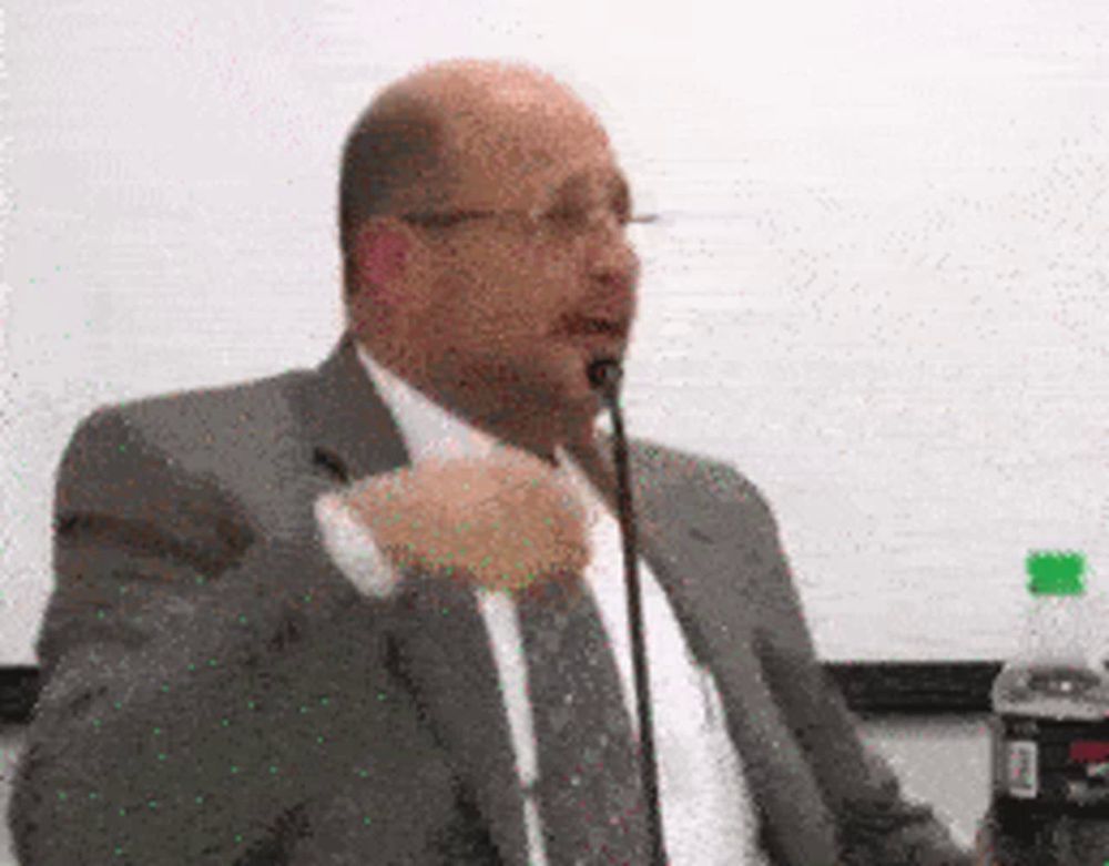 a man in a suit and tie speaking into a microphone next to a bottle of diet coke