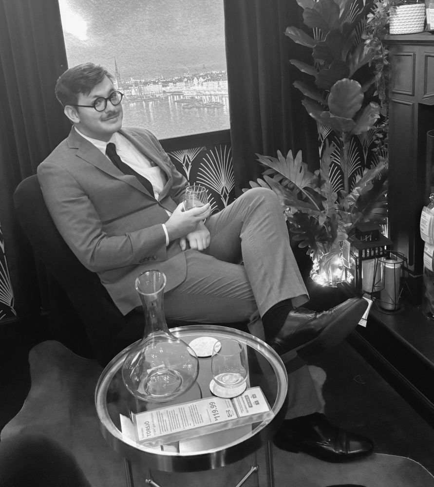 A picture of the original poster in a suit, sitting on a chair in a display in an IKEA store. The person in the photograph is wearing a gray suit and black tie, sports a mustache and has black rimmed round glasses. He is holding a glass.
