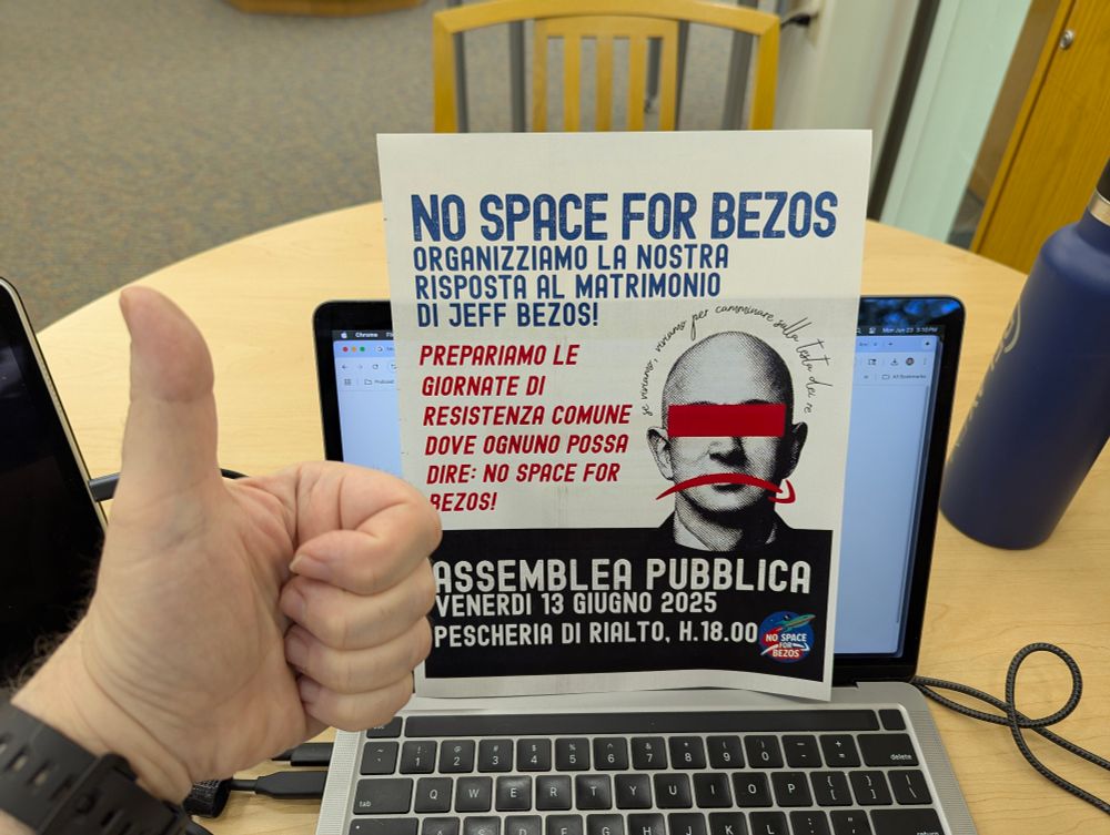 Photo of a library table. A MacBook, iPad, notepad, and water bottle are laid out as for a quiet session. A color print of a poster, printed on letter sized paper, is propped against the MacBook screen. The poster is in Italian, except for a title in English that reads: "No space for Bezos." The poster urges people of Venice, Italy to join a mass protest against the disruption Jeff Bezos' wedding is causing for the city   