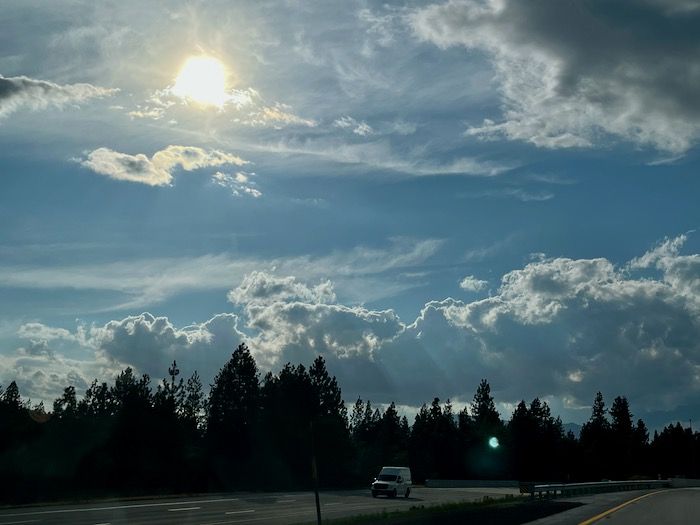 photo: the sun in the west shining through layers of wispy clouds, above other layers of chunky cloud in a grey-blue sky. Taken from highway 5 with a line of dark evergreens on the side of the road.
