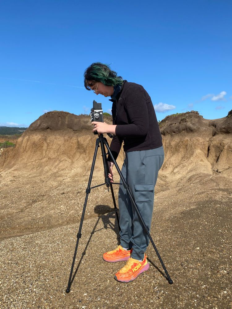 A person standing at a tripod, looking down into the camera, taking a picture of the ocean.