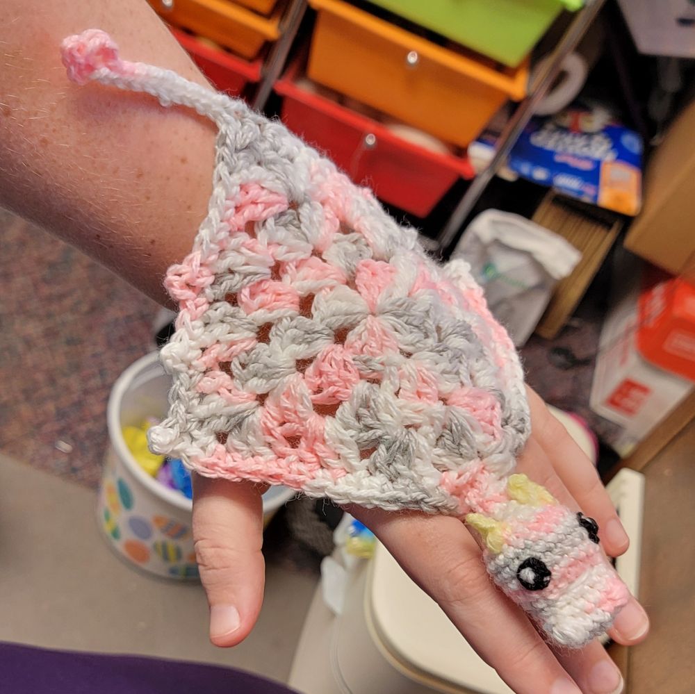 A pink, grey, and white granny square with a dragon head on the back of a hand, secured to the middle finger 
Pattern by Serendipity Designs on Etsy 