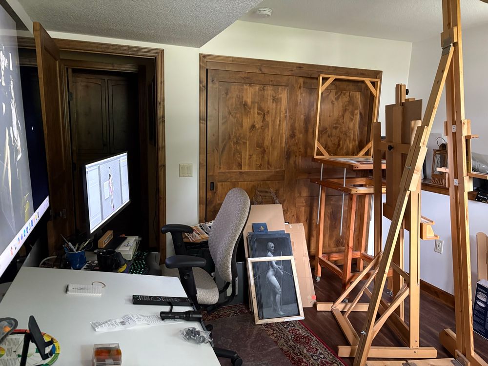 A drawing desk with computer work station beyond stack of art that’s being disappeared, an adjustable still life table and 2 easels. 