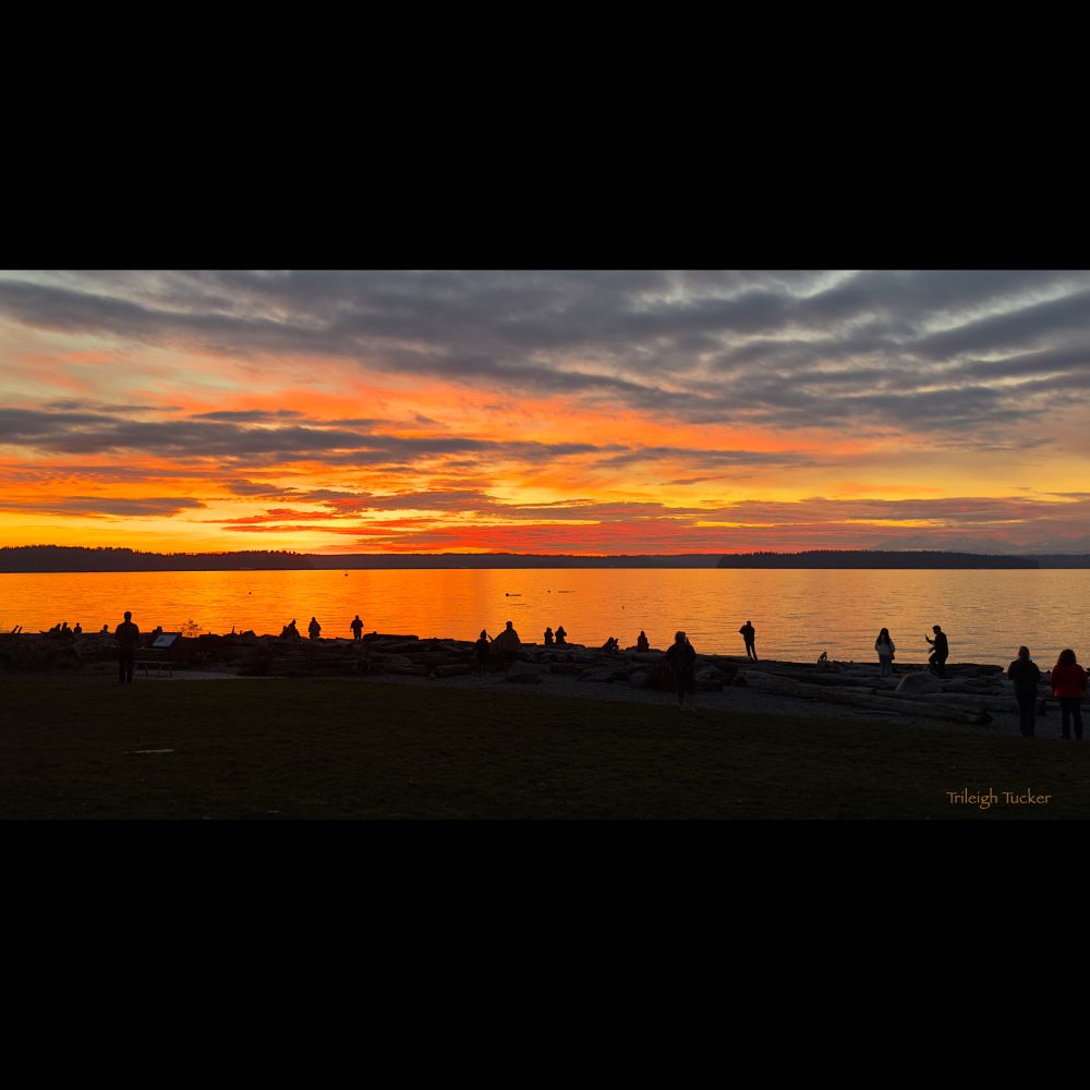 Group of people gathered at a beach, with mountains in the background, watching a colorful sunset