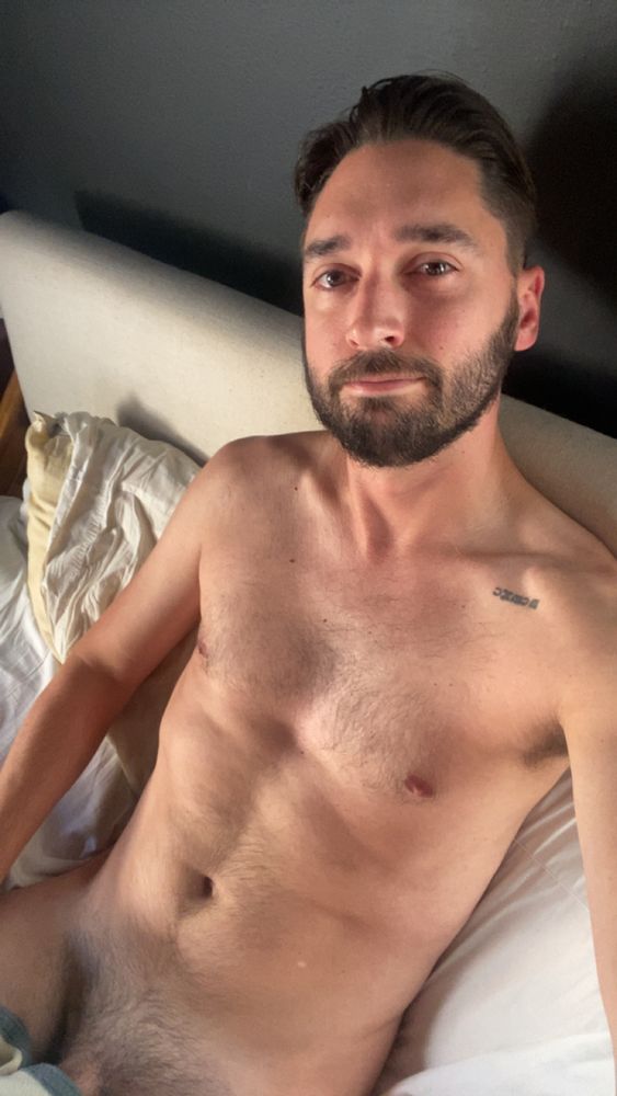 bearded user reclines in bed in the early morning light, naked down to the exposed root of his dick