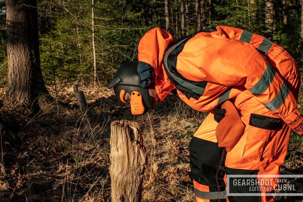Spring, a small clearing in a forest between trees, flooded with sunshine with brown leaves on the ground. Next to a knee-high tree stump stands the fox in his orange-black puppy hood, wearing his black-orange FOX MX gloves and an orange one-piece boiler suit with black shoulder straps and knee pads and reflective stripes on the lower arms, lower legs, around the belly and over the shoulders. In his right hand he holds his orange FOX cap. The camera captures the fox from the side as he leans over to sniff at the tree stump, checking that his territory is properly marked.