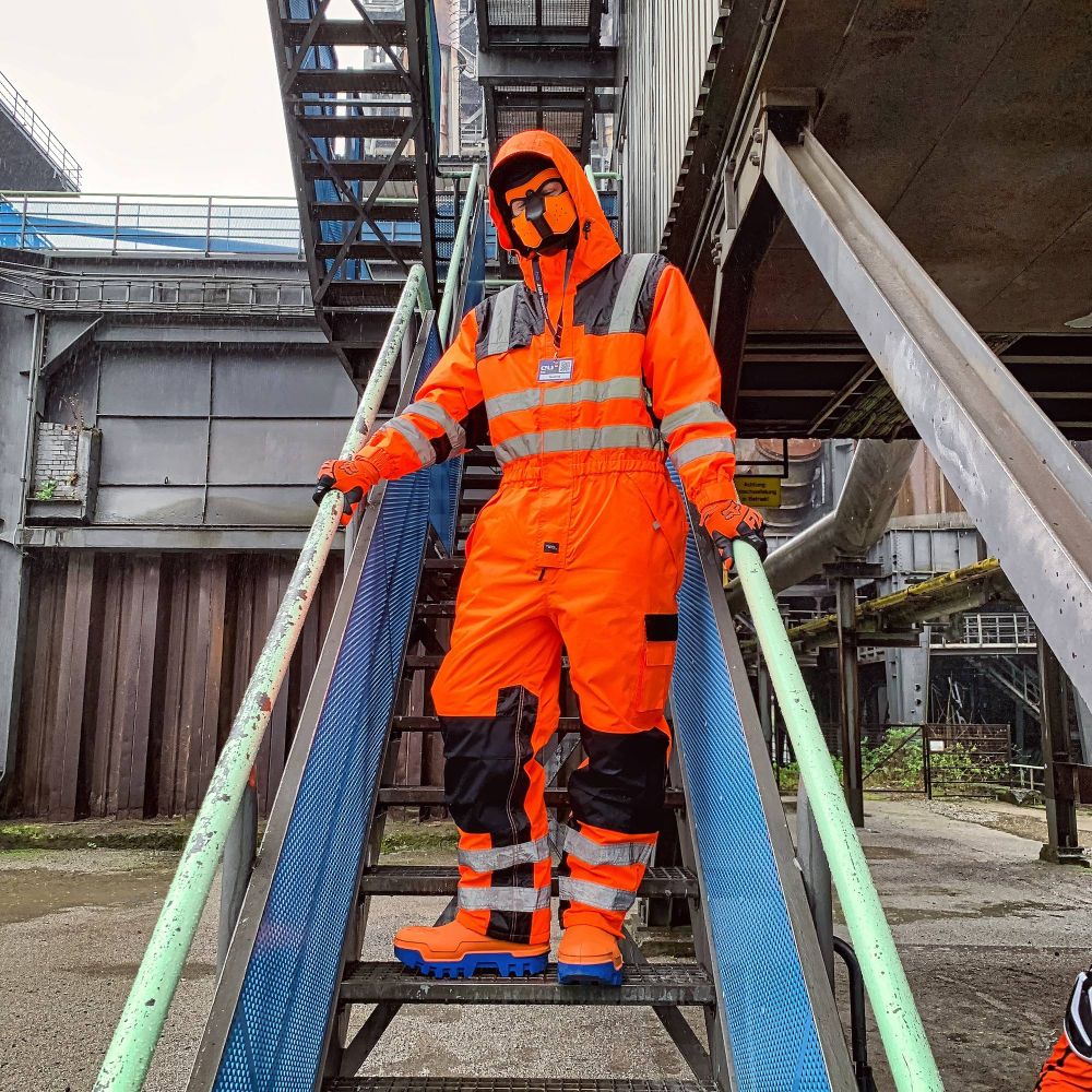 A gloomy and rainy day in an industrial areal. Several levels of an industrial complex make out the background, with metal pillars, pipes and railings. A narrow metal stairs leading up right into the complex makes out the center of the picture, with the fox standing on it, facing the camera, with his hands resting on the railing. The fox wears his orange hi-vis one-piece boilersuit with black knee and shoulder pads and horizontal reflective stripes on his lower arms, belly and lower legs, and vertical stripes over the shoulders, his black-orange puppy mask mostly covered by the orange boilersuit hood, orange-black FOX MX goves and orange rubber boots with blue soles that are mostly covered by the boilersuit.