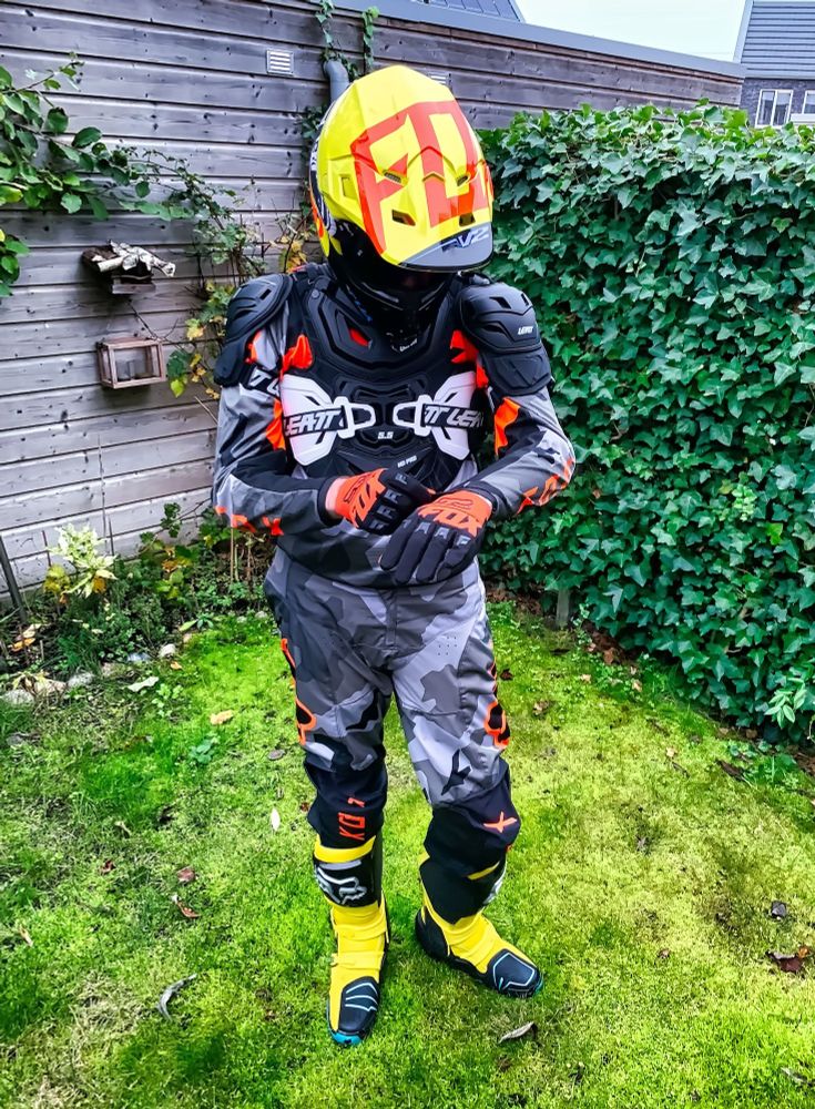 The fox stands in his full MX outfit in a garden on lush green gras. In the background is to the left a shed with plants in front of it and to the right an ivy overgrown fence that connects to the shed in a 90 degree. The fox looks down at his left hand while he's adjusting the glove of his left hand with his right hand. His full MX set consists of a grey camo-patterned FOX MX jersey with orange text highlights, orange-black FOX MX gloves, neon-yellow with blue and black highlights FOX MX boots, a black-white Leatt MX chest protector, a yellow-orange MX helmet and borderless dark-tinted MX goggles. As the fox looks down, the top side of the helmet is visible, mostly yellow, with large letters that read "FO" (from "FOX") readable in orange.