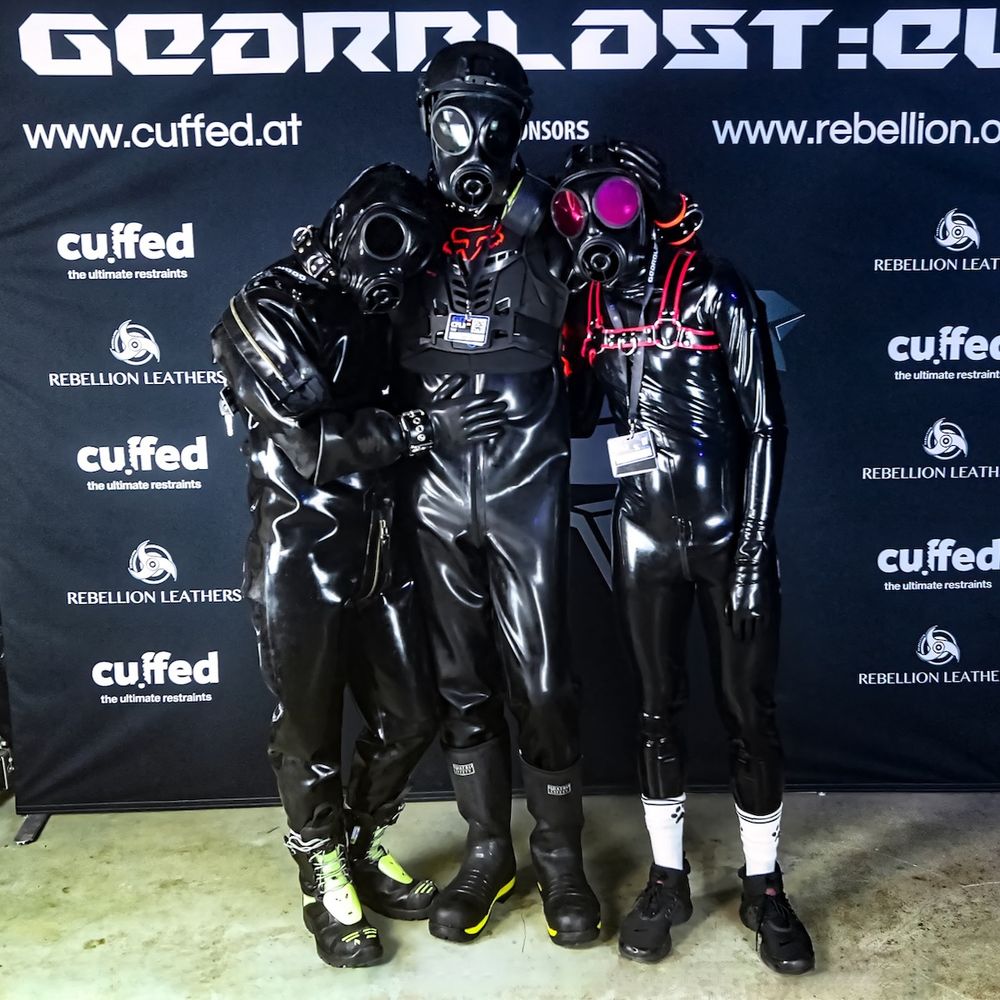 The fox drone in front of the GearBlast Europe 2022 photobox wall stand, surrounded by RedHeadGay93 and Blue Pup Milo drones to his right and left, respectively. All three are wearing rubber suits with rubber gloves, rubber hoods and S10 gas masks. While facing the camera, the fox holds both drones with his hands on their backs, with RedHeadGay93 resting his hand on the belly of the fox.
