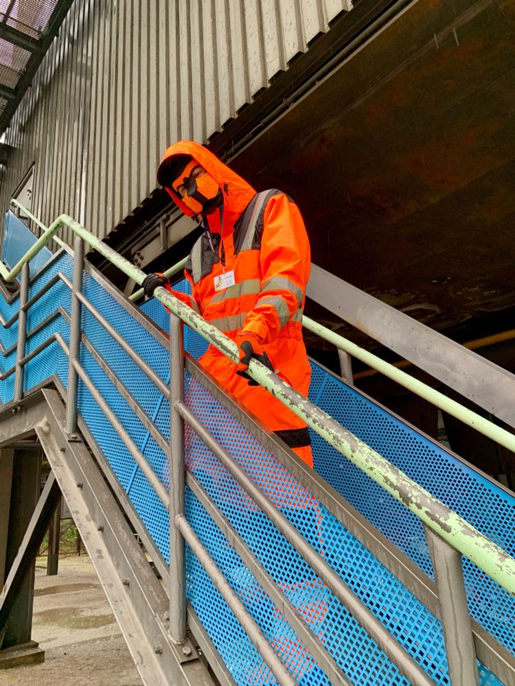 A gloomy and rainy day outside in an industrial areal. Several levels of an industrial complex make out the background. Side-shot of a narrow metal stairs leading up right into the complex, with the fox standing on it, facing down to the camera, with his hands resting on the railing. The sides of the stairs are made of a blue metal mesh, with the legs and feet of the fox barely visible through the mesh. The fox wears his orange hi-vis one-piece boilersuit with black knee and shoulder pads and horizontal reflective stripes on his lower arms, belly and lower legs, and vertical stripes over the shoulders, his black-orange puppy mask mostly covered by the orange boilersuit hood, orange-black FOX MX goves and orange rubber boots with blue soles that are mostly covered by the boilersuit.