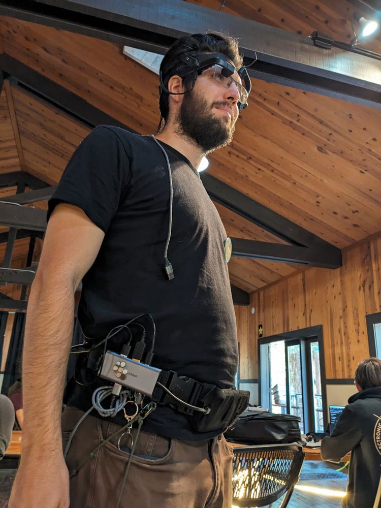 Zacchaeus standing with his full cyborg getup: heads up display, a USB-C dock, a belt, cables leading everywhere including to (out of view) ergonomic keyboards