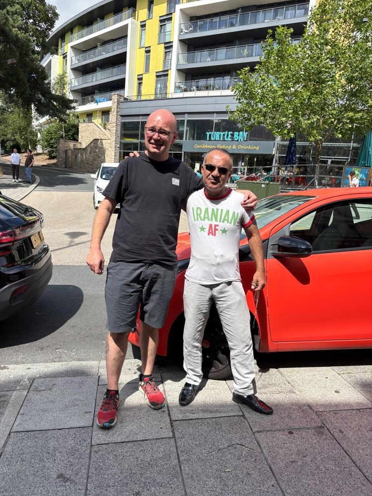 2 men starting on a pavement . one bald with glasses. The other short but very powerful wearing a t-shirt stating he is Iranian as fuck.