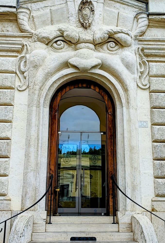 Eine imposante, helle Treppe zu einem Glasportal, in dem sich der blaue Himmel und gegenüberliegende Gebäude spiegeln. 
Das Portal ist ein weit geöffneter Mund eines Monsters – Nase, Augen und Ohren hoch über der Tür, – welches Besucher beim Eintreten zu verschlucken droht.
"Haus der Monster", 
Fassade aus dem 16.Jhd.

"House of Monsters", 
C16 facade
Imposing, light-coloured stairway leading up to a large glass portal which reflects blue sky and buildings across. 
The doorway is striking because it is the wide-open mouth of a face - nose , eyes, ears high above the door - swallowing up visitors as they enter. 