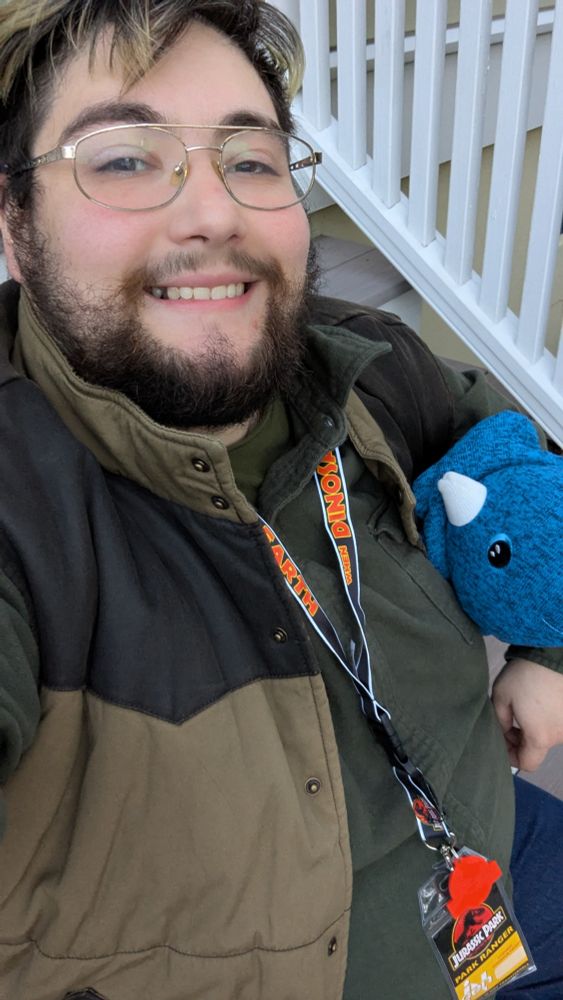 A man sitting outside dressed in a green flannel shirt and brown vest. A lanyard that says Jurassic Park ranger hangs from his neck. He is holding a blue plush triceratops 