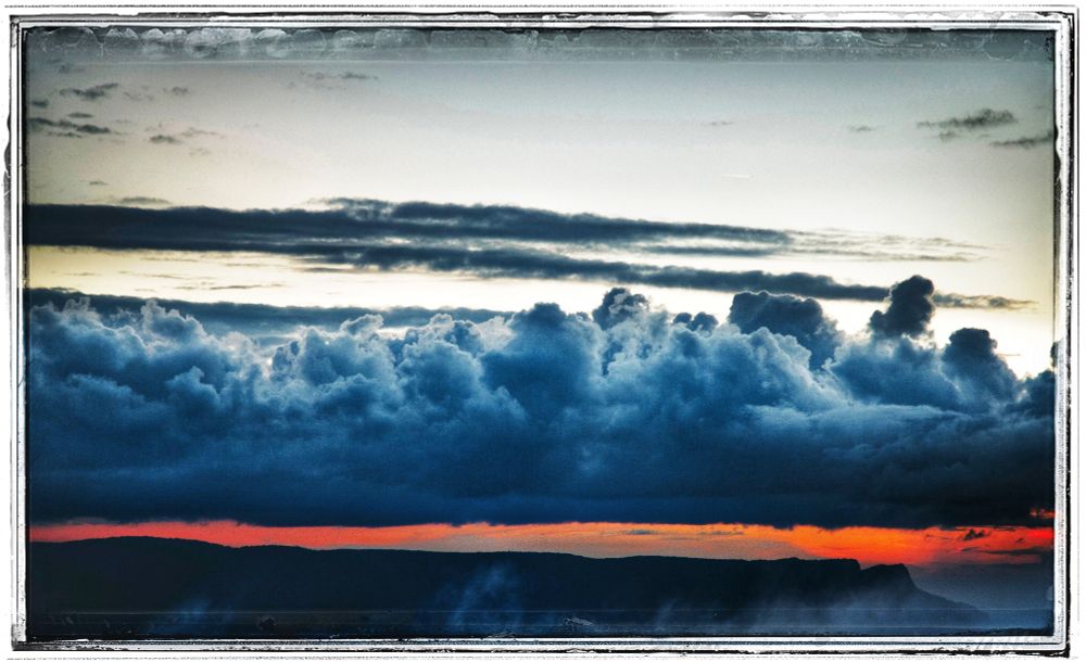 

Das Bild zeigt einen dramatischen Himmel mit mehreren Schichten von Wolken. Im unteren Bereich des Bildes befindet sich eine dünne rosarote oder orangefarbene Linie, die als Horizont oder Sonnenuntergang interpretiert werden kann. Darüber erhebt sich eine dichte Schicht dunkelblauer und grauer Wolken. 