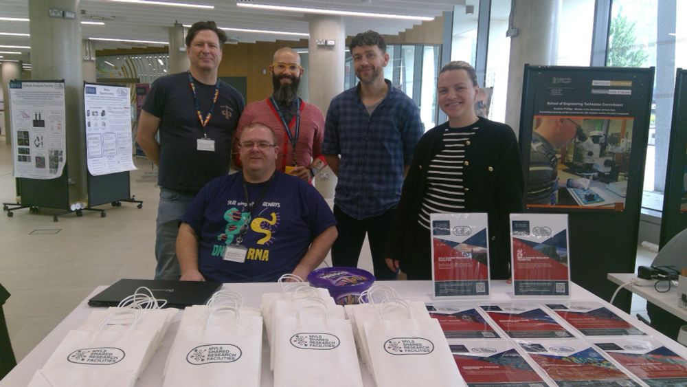 Photo of 5 people standing in front of a table with paper bags and small A4 posters.