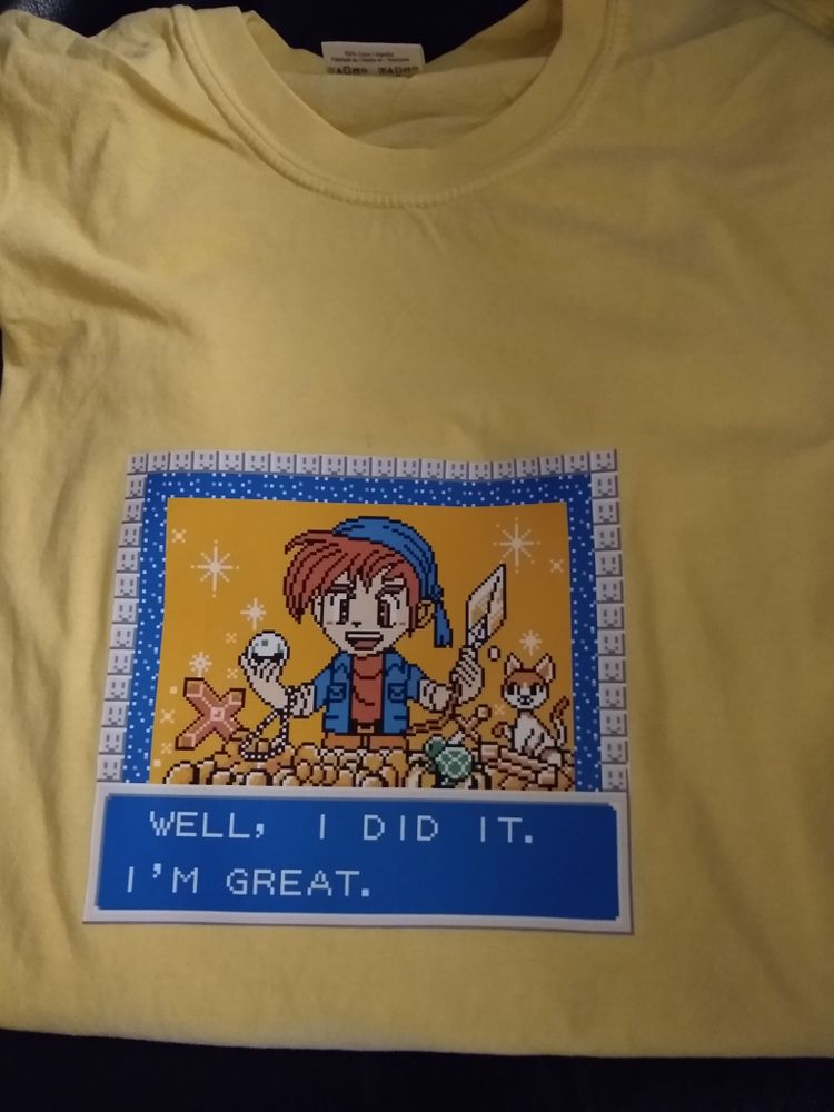 Yellow T-shirt decorated with a game screenshot showing an anime-ish adventure guy holding and beholding a pile of treasure, accompanied by a small  cat. Text box at the bottom of the screenshot reads: "WELL, I DID IT. I'M GREAT."