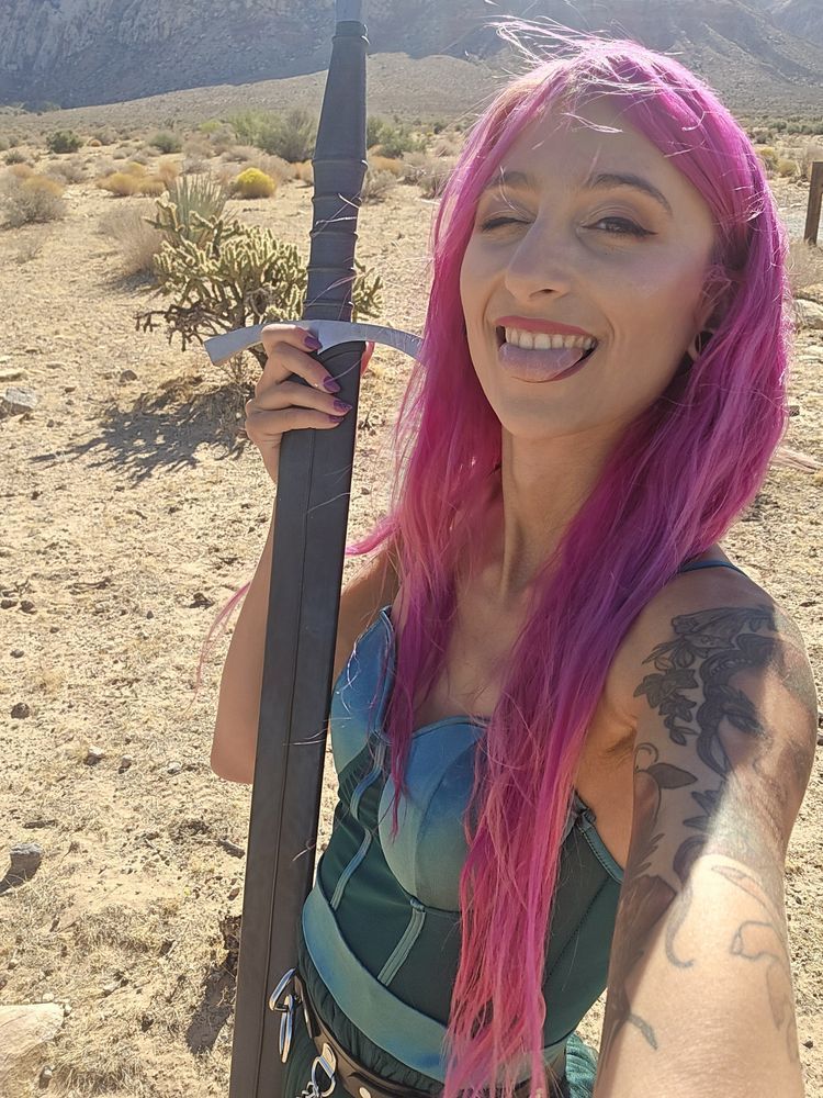 A person with long pink hair is wearing a green dress and is holding up a sheathed sword while standing in a desert. They are looking at the camera, winking, with their tongue out.