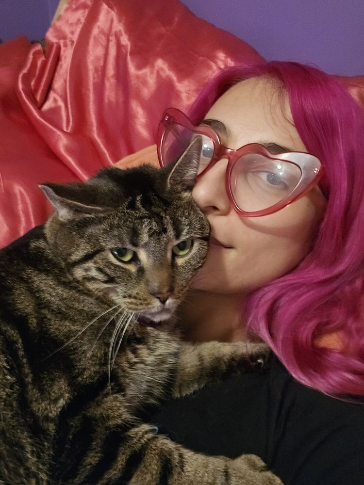 A person with long pink hair is laying in a pink bed with a tabby cat on their chest. Both are looking into the camera.