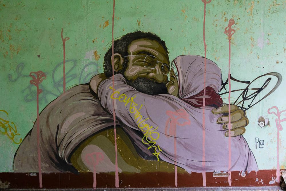 A mural by American artist Pat Perry showing the imagined release of Guantánamo captive Shaker Aamer, a large bearded man wearing glasses, hugging a woman wearing a headdress. Aamer remained in captivity while Perry was painting this in 2014 and was only released in October 2015, despite being cleared for release in 2007. He was incarcerated for 14 years altogether. He was never charged with a crime. 