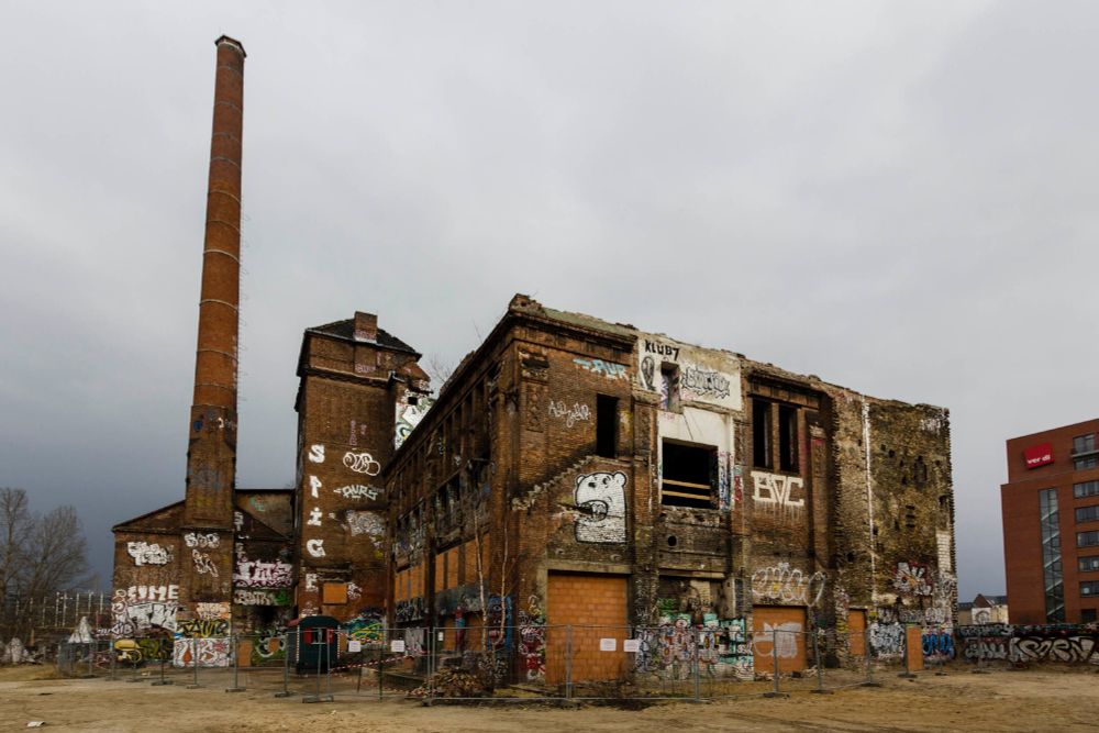 A derelict graffiti-covered squat red-brick building with a long thin chimney reaching for the grey sky behind.