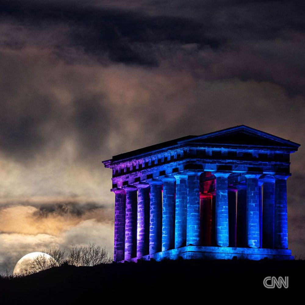 The moon sets behind Penshaw Monument in Sunderland, United Kingdom, which appears illuminated with blue, reddish and purple lighting