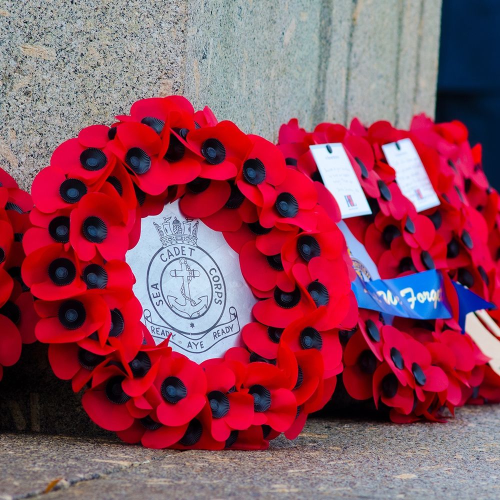 A close-up of a red poppy wreath resting against a stone memorial. The center of the wreath features the Sea Cadet Corps emblem with the motto "Ready Aye Ready."