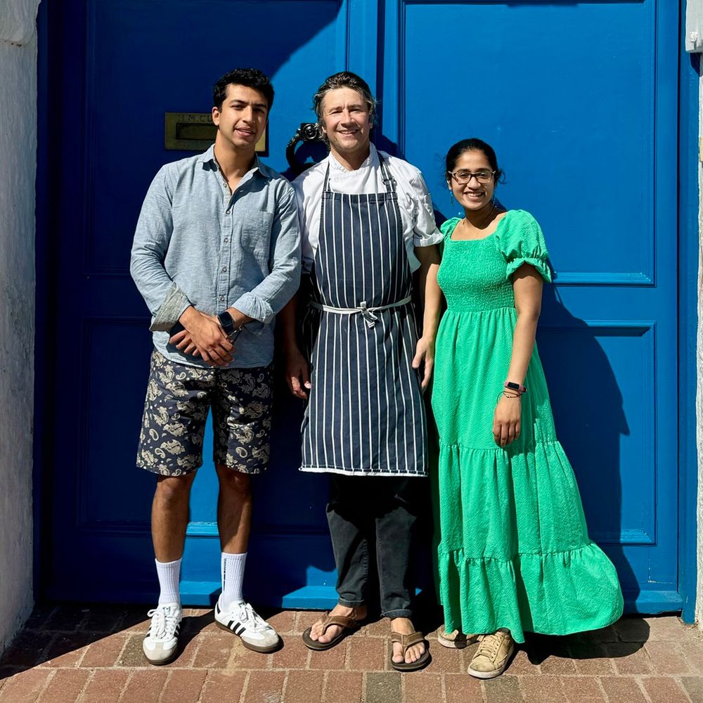 Three people standing and smiling in front of a bright blue door. The man in the middle wears a chef’s apron, while the two others—one in a green dress and the other in a blue shirt with patterned shorts—stand on either side.