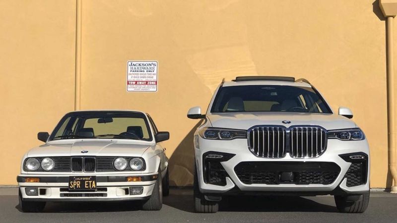An old E30 series BMW looking like a toy car next to a modern X7 from 4 decades later. The bonnet of the SUV is about at roof height of the old sedan. 