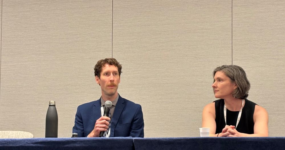 A man in a blue suit speaks with a microphone while a woman in a sleeveless black shirt looks on. 