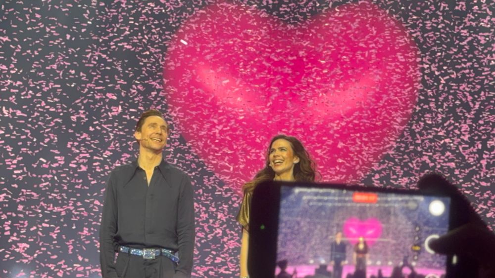 Pink confetti is falling there is a giant red inflated heart in the back Tom Hiddleston and Hayley Atwell are smiling at the audience while standing next to one another on stage.