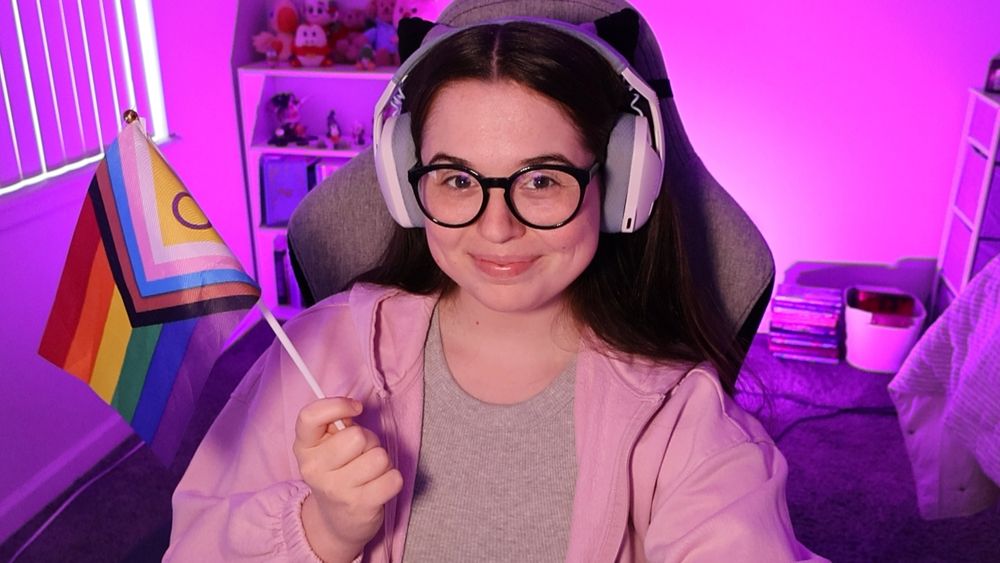 selfie of kate holding up the intersex pride progress flag and smiling at the camera. they're wearing a grey tank top with a light pink zip up hoodie over it. they're also wearing dark blue glasses and blue and white headphones.