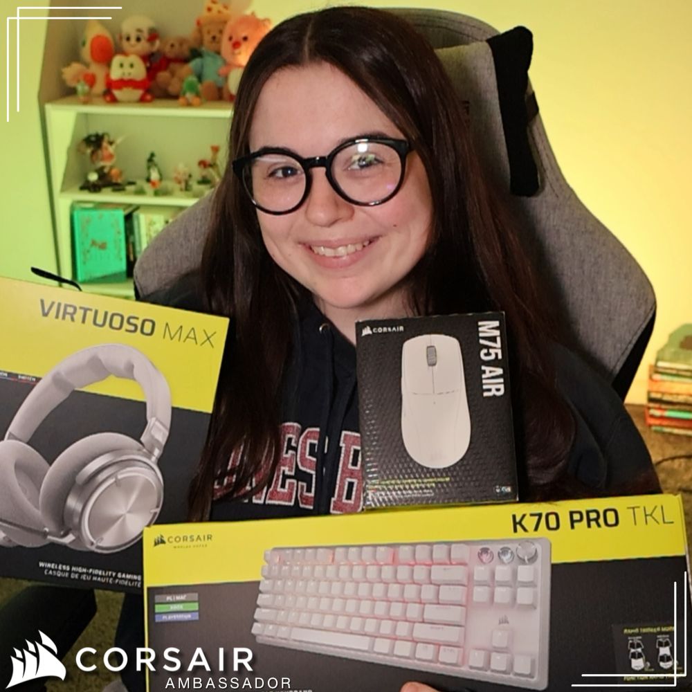 picture of kate smiling and holding up three corsair products: their virtuoso max headset, the k70 pro tkl keyboard, and the m75 air wireless mouse. they're wearing a dark blue hoodie and dark blue glasses. the background is lit up a bright yellow color. the words "corsair ambassador" are put in the bottom left corner.