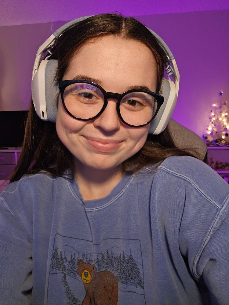 selfie of Kate smiling. they're wearing a blue sweatshirt with a bear on it, dark blue glasses, and blue and white headphones.