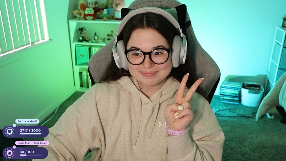selfie of kate smiling and holding up a peace sign. they're wearing a beige hoodie, dark blue glasses, and silver headphones.