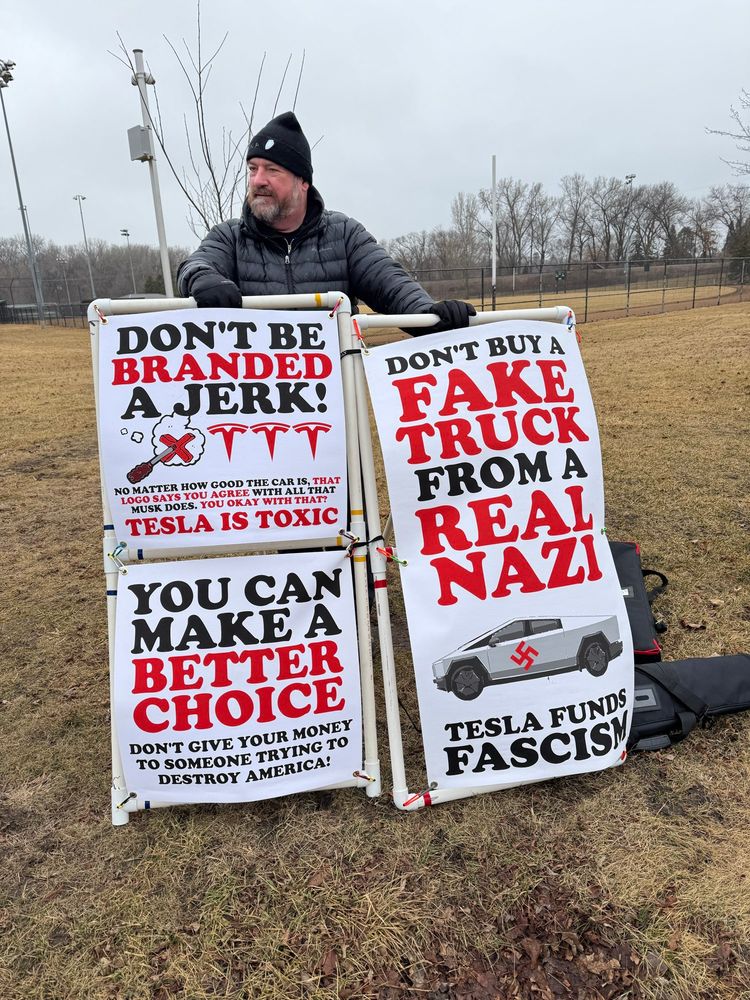 A man stands behind multiple signs, protesting, Tesla and Elon Musk. One of them says “ don’t buy a fake truck from a real Nazi. Tesla funds fascism.”