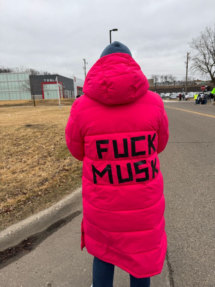A person is standing with their back to the camera, wearing a long pink winter coat with the words “Fuck Musk” on the back, spelled out with black tape. This is outside of a Tesla dealership to protest Elon Musk in Golden Valley, Minnesota.