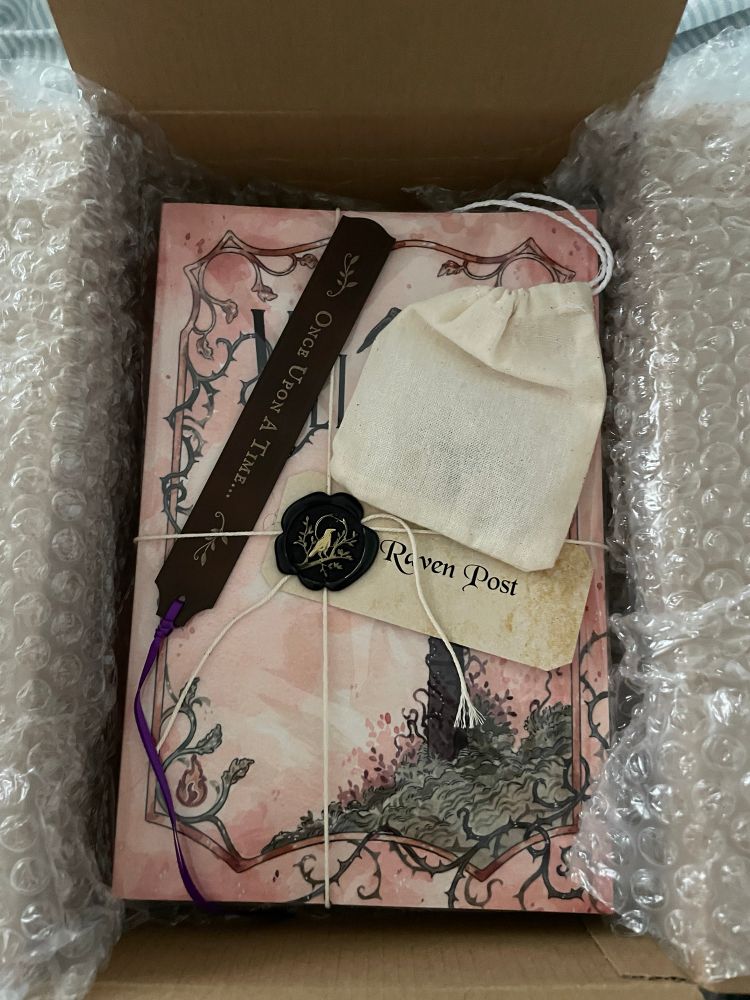 An open box containing a peach-colored book tied with string. A label marked “Raven Post” is attached to the string with black sealing wax stamped with a raven in gold. Tucked under the string is a thin black bookmark with a purple ribbon and the words “Once upon a time …”. A small muslin drawstring bag lies nearby.