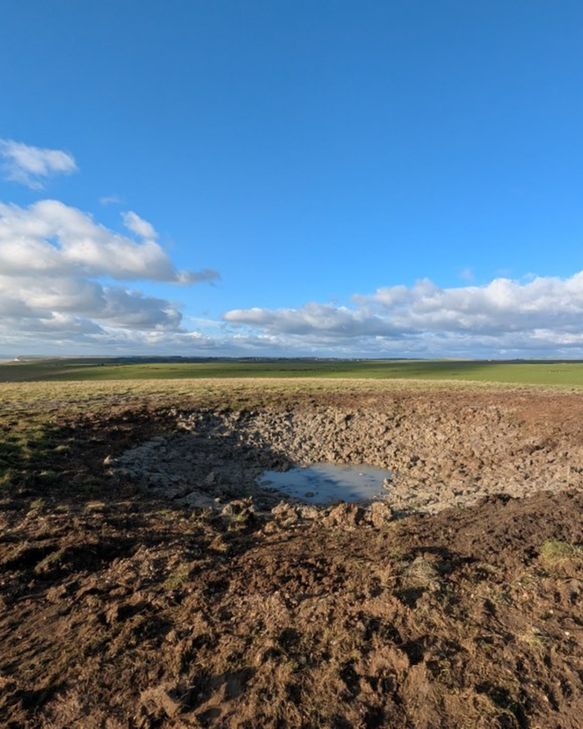 An empty dew pond under a blue sky. There's exposed mud and a small puddle in the middle. 