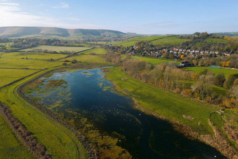 A drone image of a large stream within a landscape of rolling green hills. To the right of the stream is a small settlement with houses nestled inbetween trees. 