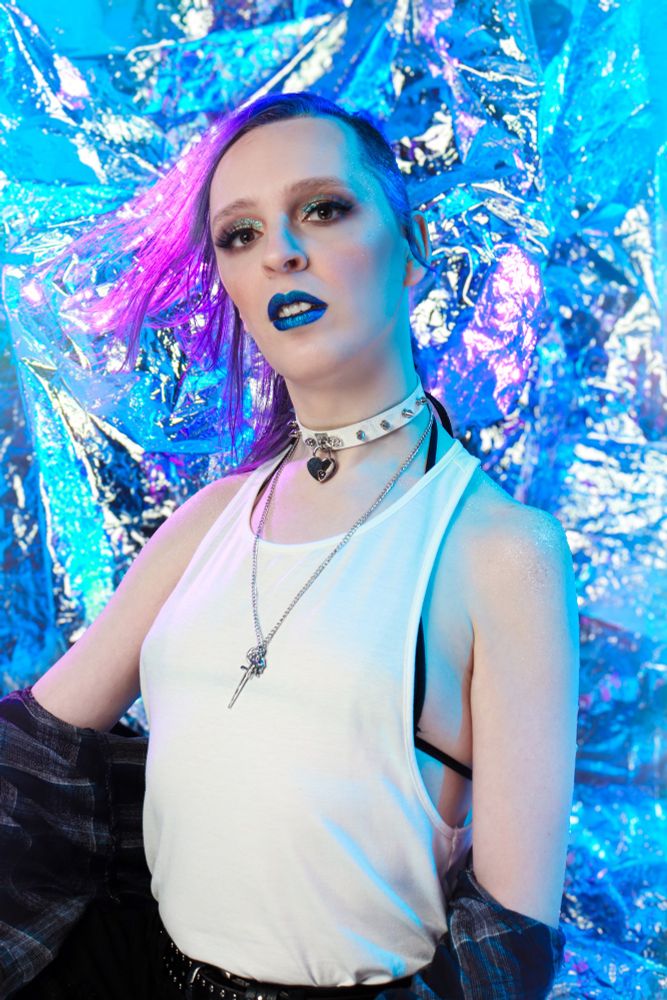 A transfemme in a white tank top, black string bikini top, a blue flannel button-up, and ripped black jeans. She's wearing blue lipstick. Her hair is a bright purple. She's looking at the camera, hair windswept.