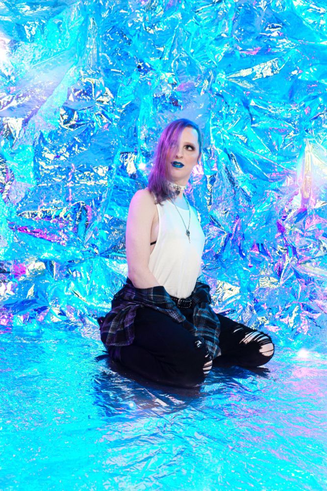 A transfemme in a white tank top, black string bikini top, a blue flannel button-up, and ripped black jeans. She's wearing blue lipstick. Her hair is a bright purple. She's looking up and to the side with a small smirk.