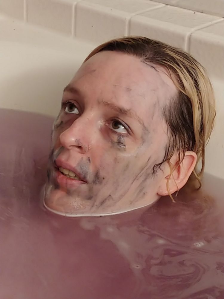 A transfemme laying in a tub, submerged up to the chin. The water is purple, their face is streaked with black stains. They look kinda dead.