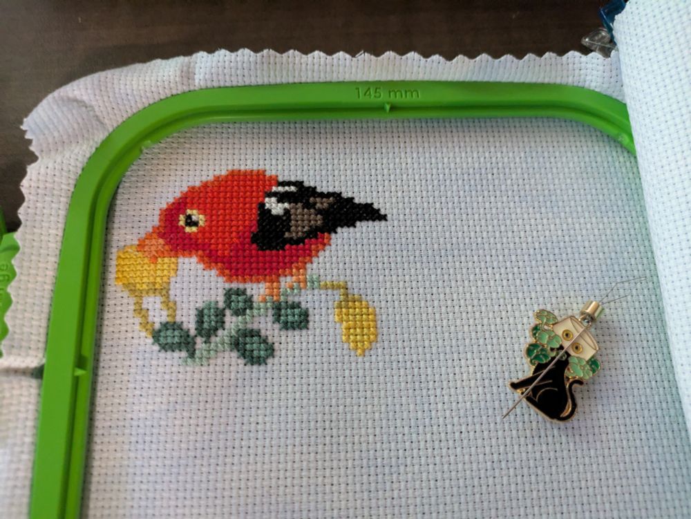 A cross stitched piece featuring an 'I'iwi, a scarlet colored bird. This one is sticking its long curved beak into a flower. 