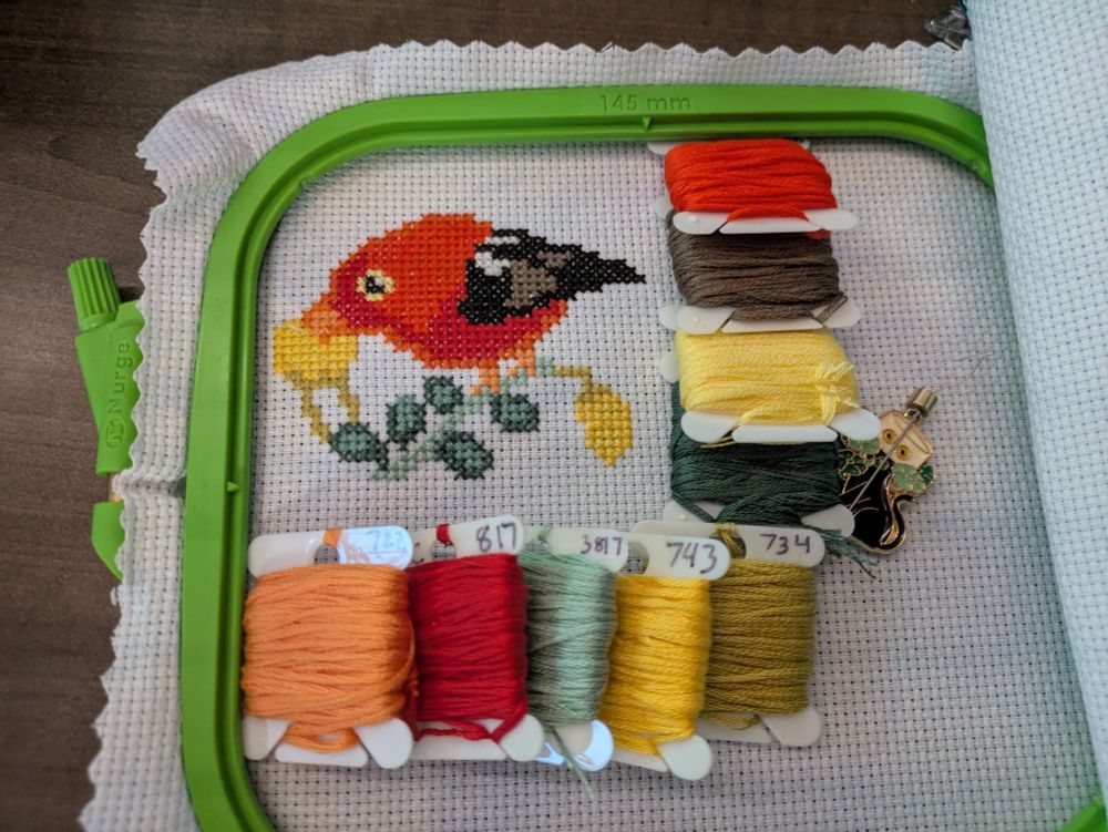 A cross stitched piece featuring an 'I'iwi with some thread bobbins framing it.