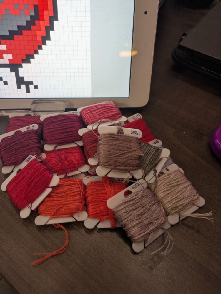 A pile of various red and gray embroidery thread in front of a pixel art doodle. 