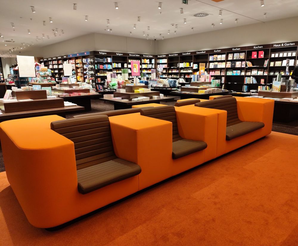 Foto der neu eröffneten Buchhandlung Rupprecht in Erlangen. Im Hintergrund Bücherregale, vorne sehr komfortabel aussehende Sitzgelegenheiten: breite Sitze mit Lehne in braun, je links und recht breite Flächen daneben zum Ablegen von Büchern oder den eigenen Armen in kräftigem orange. 