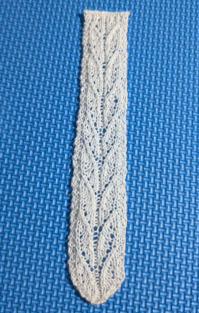 A knit lace bookmark made with white glittery thread with leaf like lace patterns going down the center on top of a bright blue background 