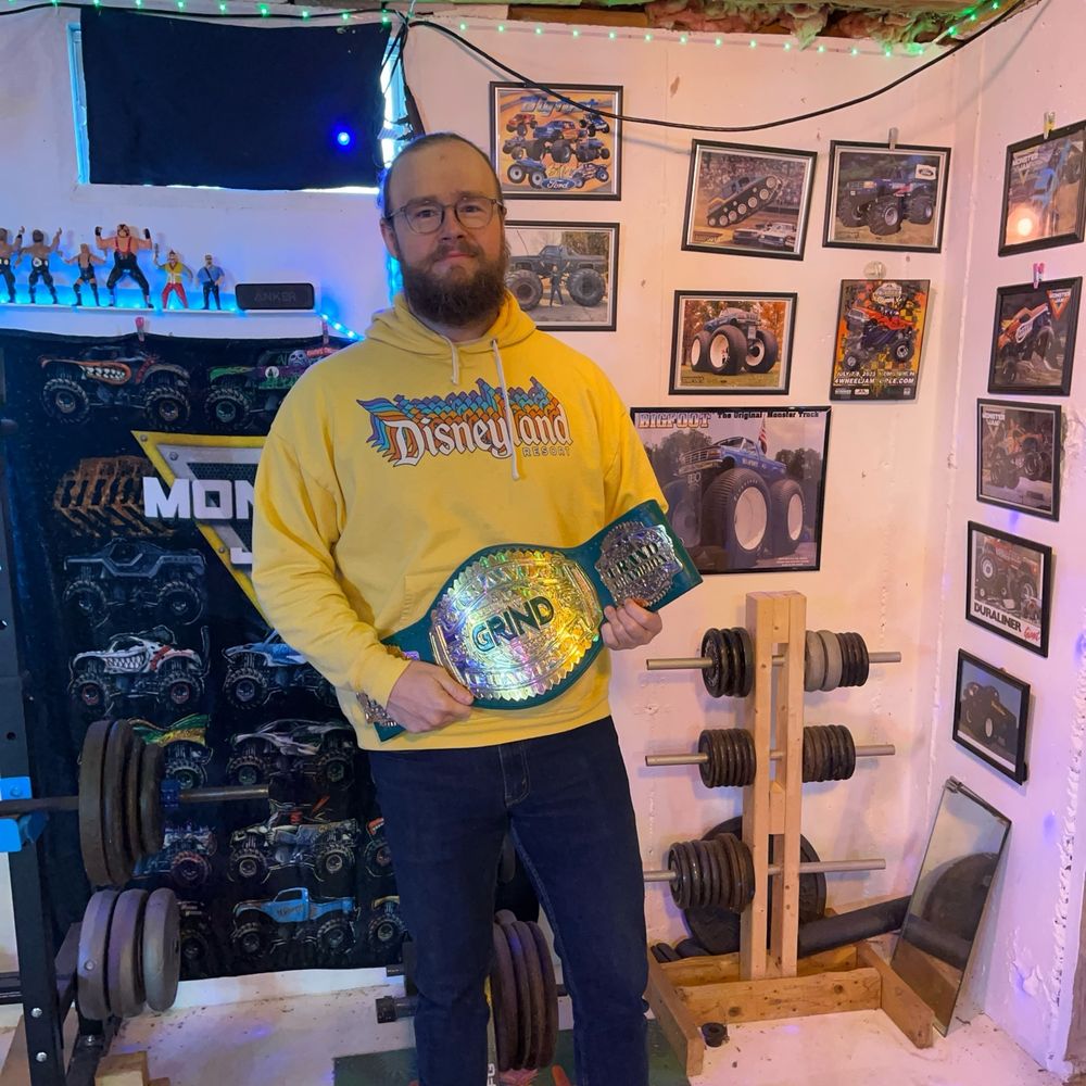Perry Von Vicious holding his newly-won Pro Wrestling Grind Grand Championship in his basement gym, surrounded by monster truck photos, action figures, and wearing a Disneyland hoodie.